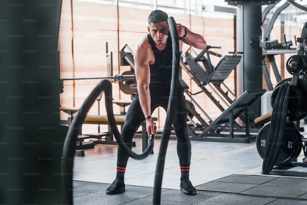 Exercising with knots. Young sportive strong man in black wear have workout day in gym.