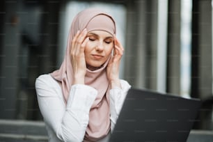 Arabian woman in hijab sitting on stairs with modern laptop on knees and touching her temples because of strong headache. Female freelancer with closed eyes suffering from overwork.