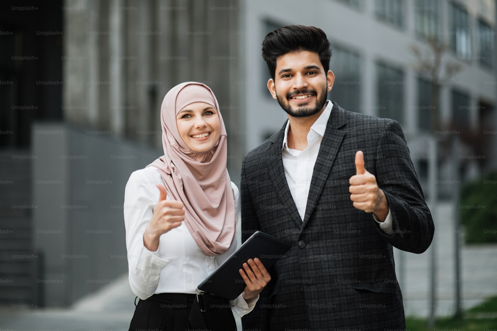 Smiling woman in hijab and muslim handsome man showing thumbs up while standing outdoors near office building. Successful cooperation of two business partners.