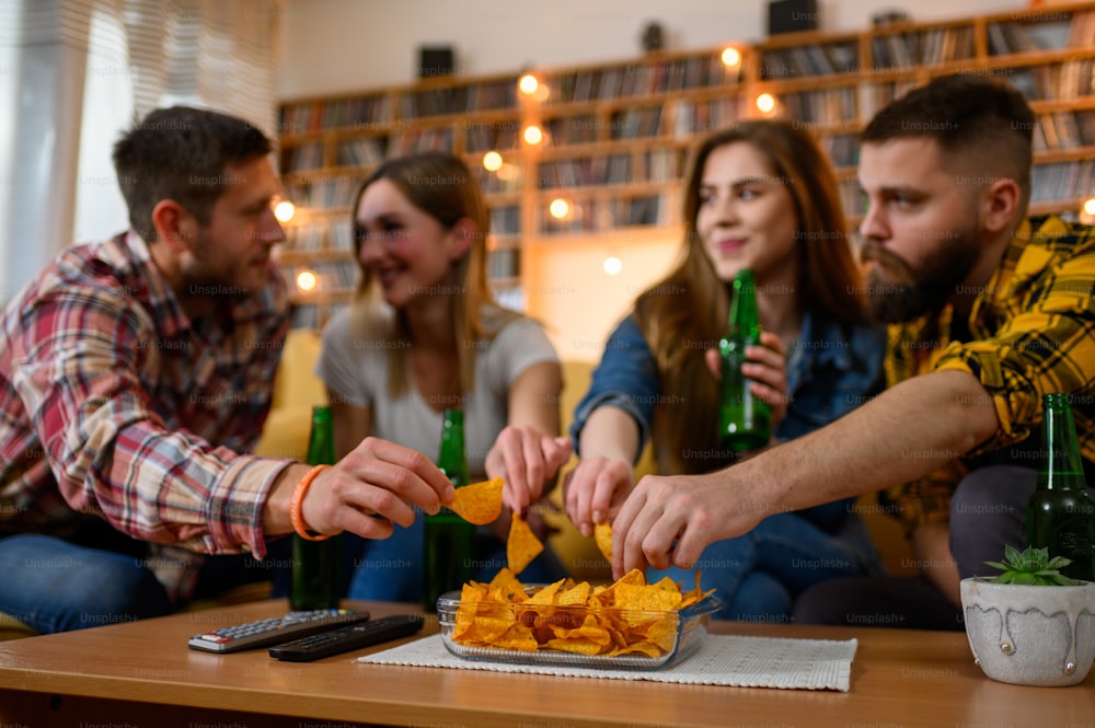 Group of friends on a house party drinking beer and eating nachos while having fun and sitting on a couch