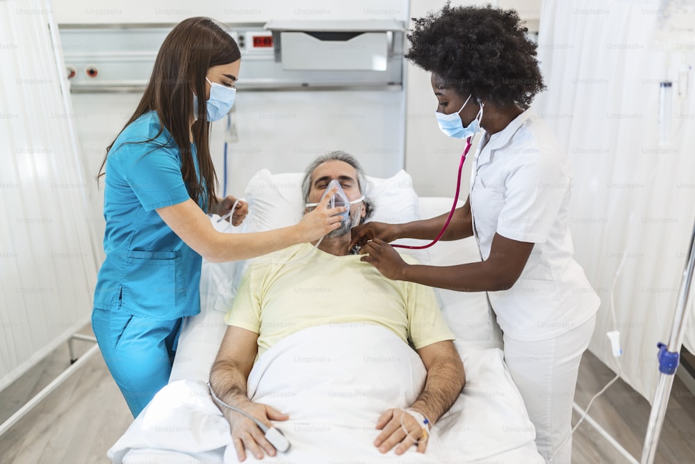 Un joven médico y una enfermera con una mascarilla quirúrgica controlan a un paciente masculino mayor que lleva una mascarilla de oxígeno de presión positiva para ayudar a respirar en una cama de hospital durante la pandemia de Covid-19
