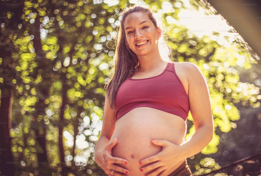 Smiling pregnant woman in sport cloth standing in the park and touching her belly.