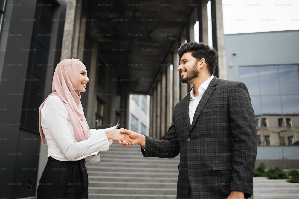 Geschäftsmann, Frau im Hijab und muslimischer Geschäftsmann, die sich die Hände schütteln, während sie draußen in der Nähe eines modernen Bürozentrums stehen. Konzept der Zusammenarbeit und des Erfolgs.