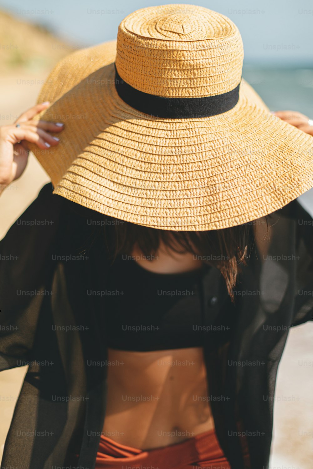帽子をかぶった美しいのんきな女性が海の波で砂浜を歩き、リラックスして、クローズアップします。夏休み。熱帯の休暇を楽しんでいる明るい黒のシャツと麦わら帽子のスタイリッシュフィットの若い女性