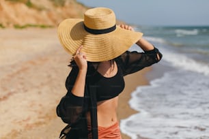 Stylish carefree woman in hat walking on sunny beach at sea and enjoying summer vacation. Young female in straw hat and light shirt relaxing on tropical island. Summer vacation
