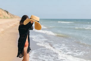 Carefree happy woman with windy hair holding hat and having fun on sandy beach at sea. Summer vacation. Stylish fit young female in light shirt and straw hat relaxing on tropical island