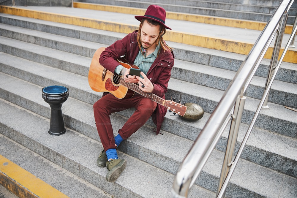 Junger Straßenmusiker sitzt auf einer Treppe, während er vor dem Auftritt auf den Bildschirm seines Gadgets schaut