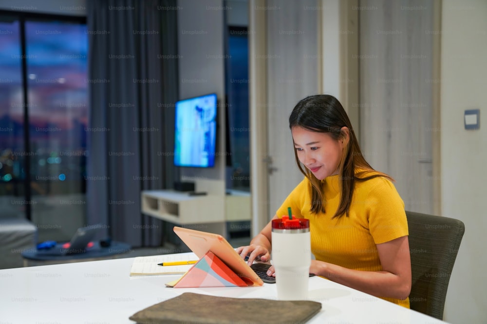 Mulher asiática bonita usando tablet digital com internet para o trabalho de pequenas empresas no apartamento à noite. Mulher freelance digitando no teclado do tablet para trabalho on-line trabalho de negócios ou compras em casa.