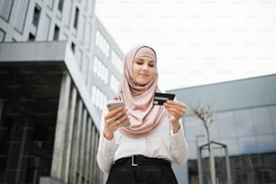 Hermosa mujer musulmana con ropa formal y hiyab haciendo compras en línea con tarjeta de crédito y teléfono inteligente. Joven de pie cerca del edificio de oficinas y disfrutando de un pago fácil.