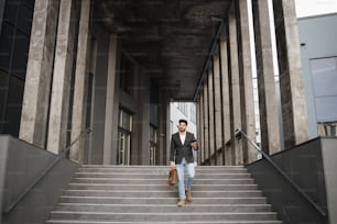 Retrato de corpo inteiro do homem árabe descendo escadas com smartphone moderno em uma mão e mala marrom de couro em outra. Conceito de negócio, sucesso e confiança.