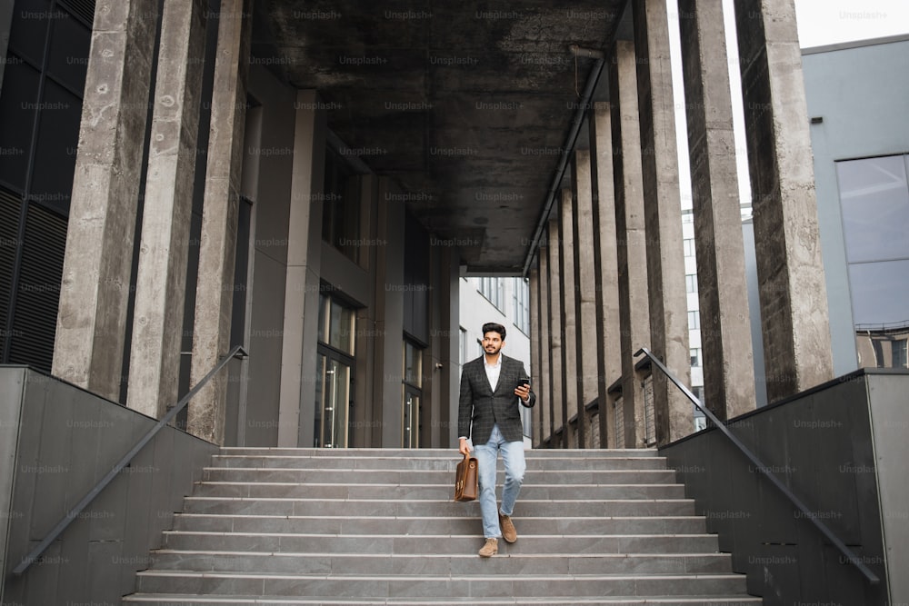 Retrato de corpo inteiro do homem árabe descendo escadas com smartphone moderno em uma mão e mala marrom de couro em outra. Conceito de negócio, sucesso e confiança.