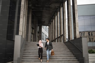 Ganzkörperporträt einer muslimischen Frau und eines Mannes, die zusammen in der Nähe eines modernen Bürogebäudes spazieren gehen und einige Arbeitsmomente besprechen. Zwei Mitarbeiter mit Smartphone, Klemmbrett und Koffer im Freien.
