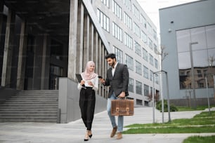 Colleghi d'affari arabi in abiti formali che camminano per strada e discutono di alcuni problemi di lavoro. Giovane uomo con valigia usando smartphone, donna in hijab che porta appunti.