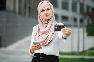 Mujer atractiva con hiyab y ropa formal sosteniendo un teléfono inteligente moderno y una tarjeta de crédito en las manos. Concepto de personas, tecnología y compra online.