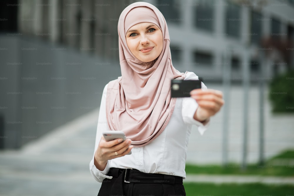 Femme séduisante en hijab et vêtements formels tenant un smartphone moderne et une carte de crédit dans les mains. Concept de personnes, de technologie et d’achats en ligne.