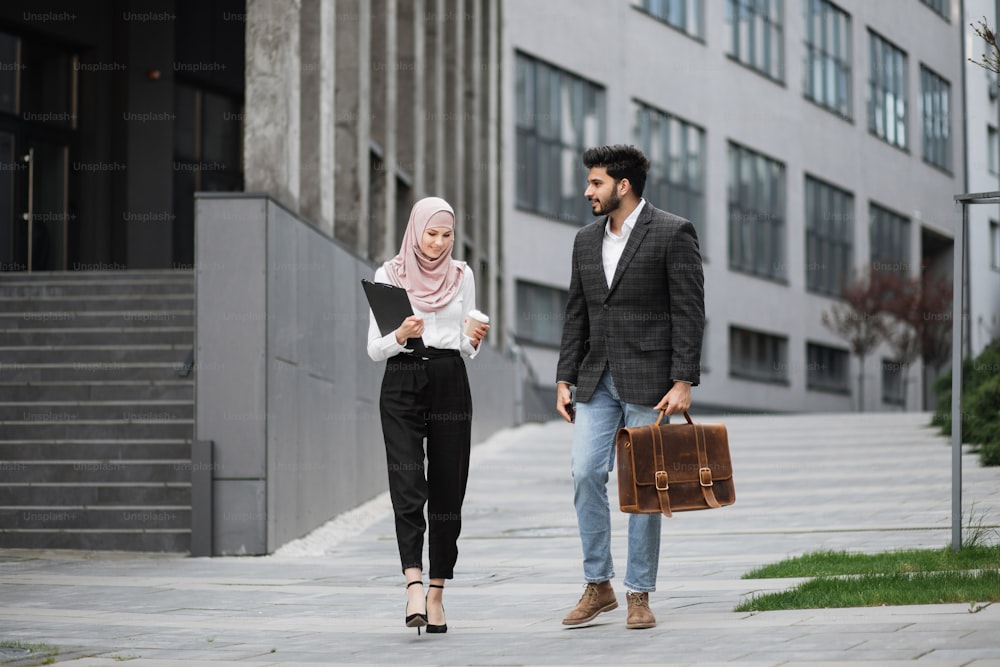 Two muslim office workers walking together outdoors smiling and chatting. Handsome man in suit carrying suitcase and smartphone, charming woman in hijab holding cup of coffee and clipboard.
