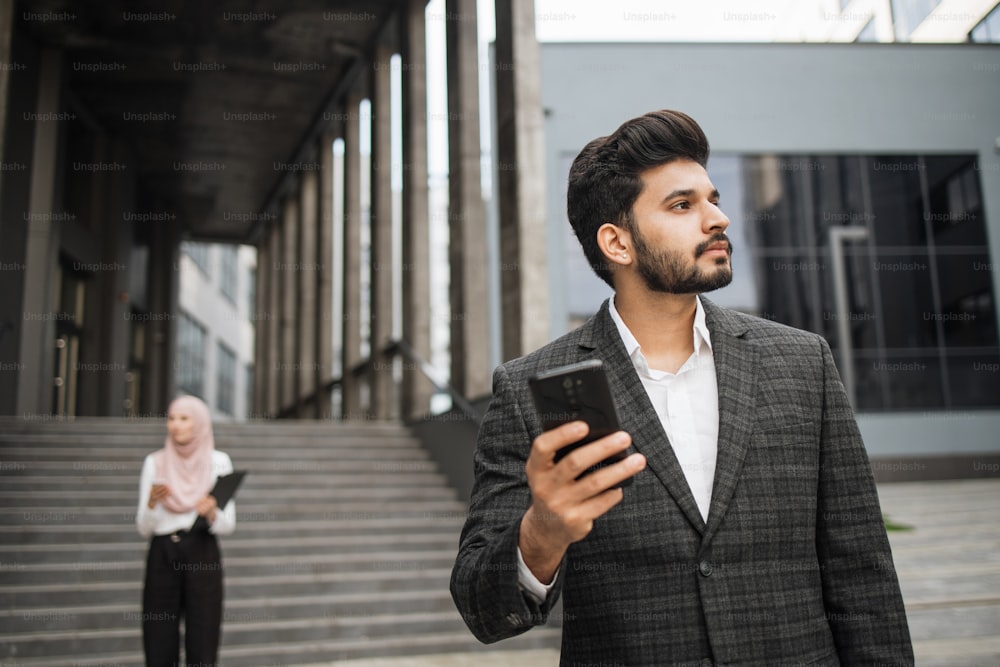 Seitenansicht eines gutaussehenden Hindu-Mannes, der Smartphone und Koffer im Freien hält. Attraktive Frau im Hijab auf dem Hintergrund mit Klemmbrett. Zwei Geschäftspartner im Freien.