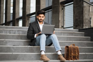 Handsome arabian man in stylish wear sitting on stairs and working on wireless laptop. Concept of people, freelance and technology.