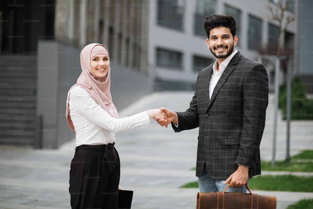 Happy arabian man and woman shaking hands after successful cooperation while standing near modern office building. Concept of people and teamwork.