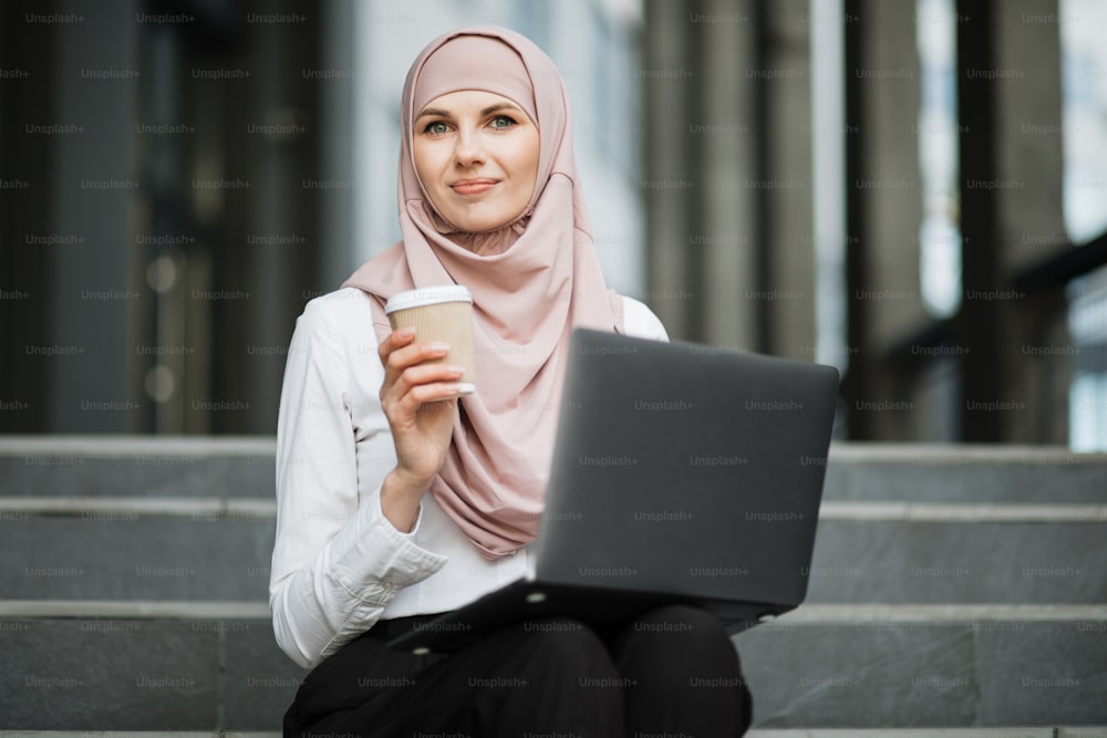 Portrait of adorable woman in hijab and elegant clothes enjoying takeaway coffee and working on portable computer outdoors. Concept of remote work and modern gadgets.