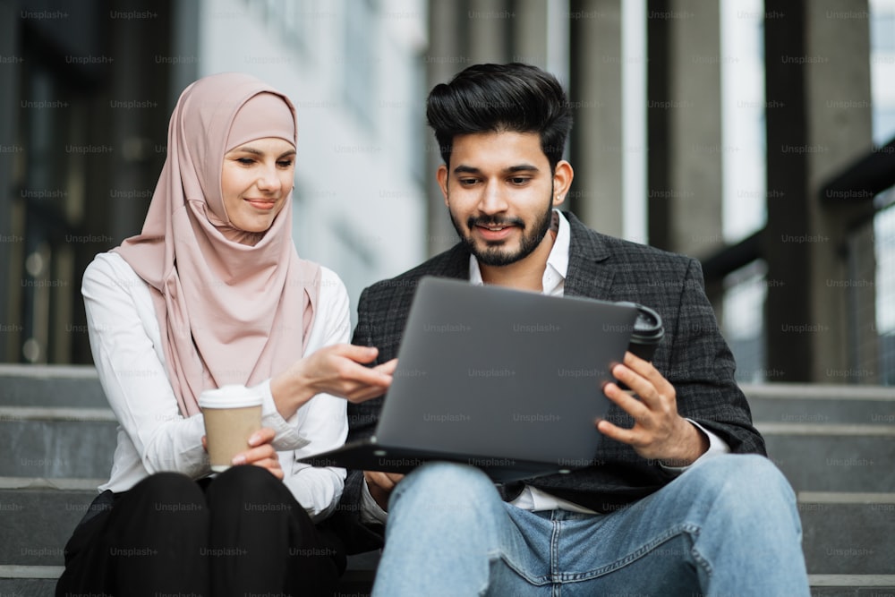 Stylish muslim man and charming woman in hijab sitting together on stairs, smiling and talking. Two freelancers using laptop for for work outdoors.