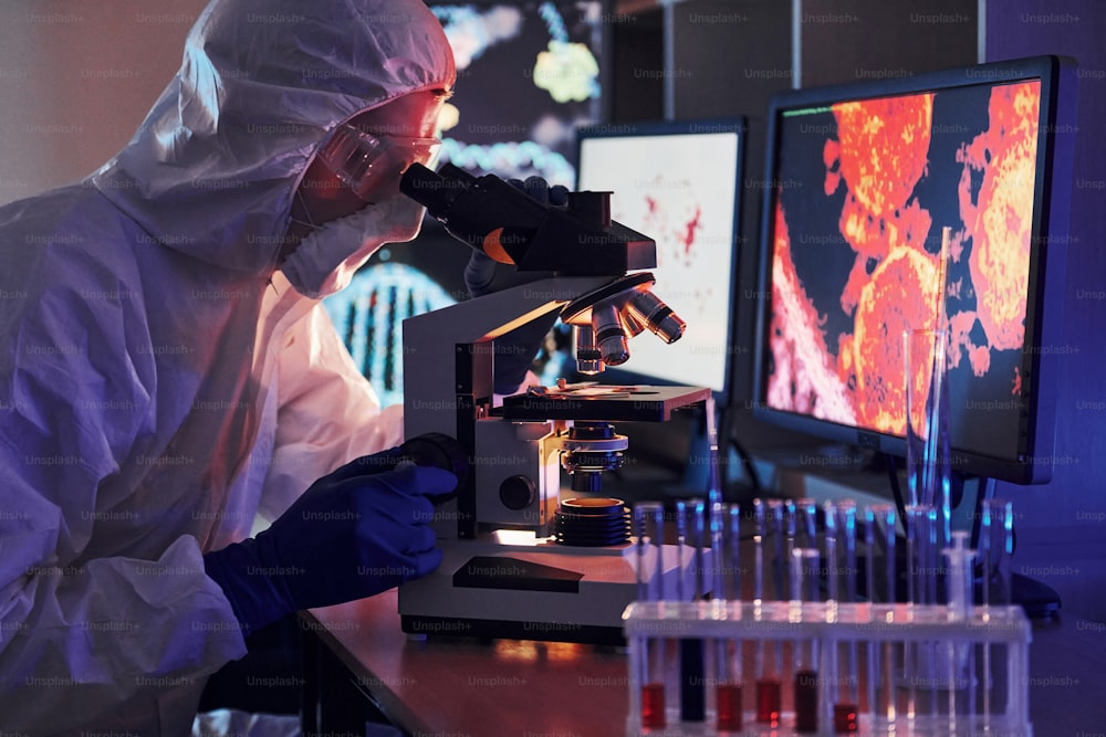 Monitores con información sobre la mesa. Un científico con uniforme protector blanco trabaja con coronavirus y tubos de sangre en el laboratorio.
