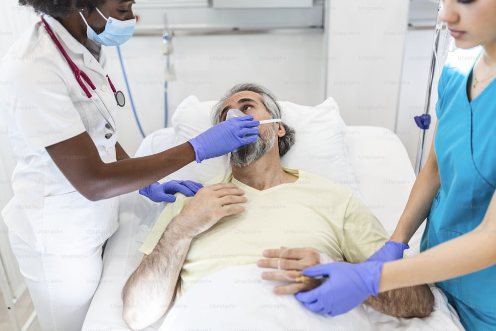 Female doctor talking with patient along coworker in ICU. Man is lying on bed amidst essential workers. Healthcare workers are in protective workwear.