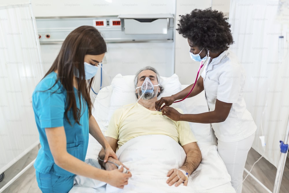 I medici con maschere protettive stanno esaminando il paziente anziano infetto in ospedale. In ospedale il paziente anziano riposa con la maschera dell'ossigeno, sdraiato sul letto.