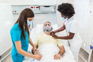 防護マスクを着用した医師が、感染した高齢の患者を病院で診察しています。病院では、高齢の患者が酸素マスクを着けてベッドに横たわって休んでいます。