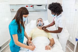 Doctors with protective masks are examining the infected aging patient in the hospital. In the Hospital Senior Patient Rests with oxygen mask, Lying on the Bed.