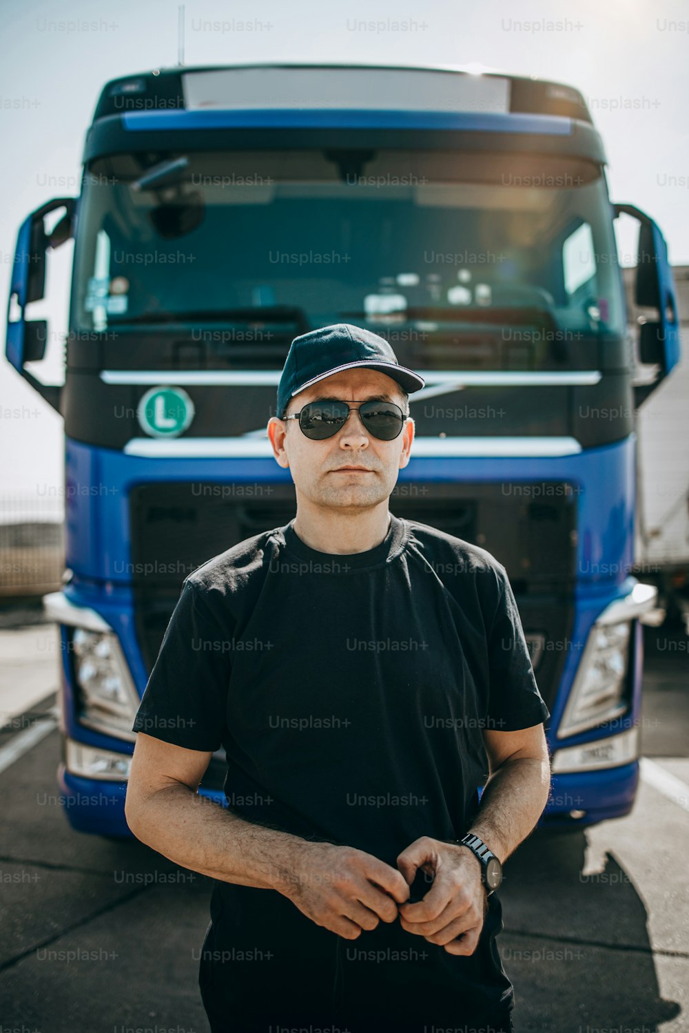 모자와 선글라스를 쓴 전문 트럭 운전사가 크고 현대적인 트럭 앞에 자신 있게 서 있습니다. 밝고 화창한 날. 사람과 교통 개념.