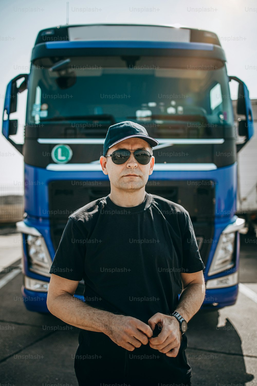 Professional truck driver with hat and sunglasses confidently standing in front of big and modern truck. Bright sunny day. People and transportation concept.