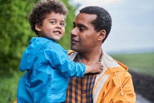 Portrait view of the happy multiracial father expressing fondness to his kid while holding him at the hands and feeling glad and tender. Stock photo