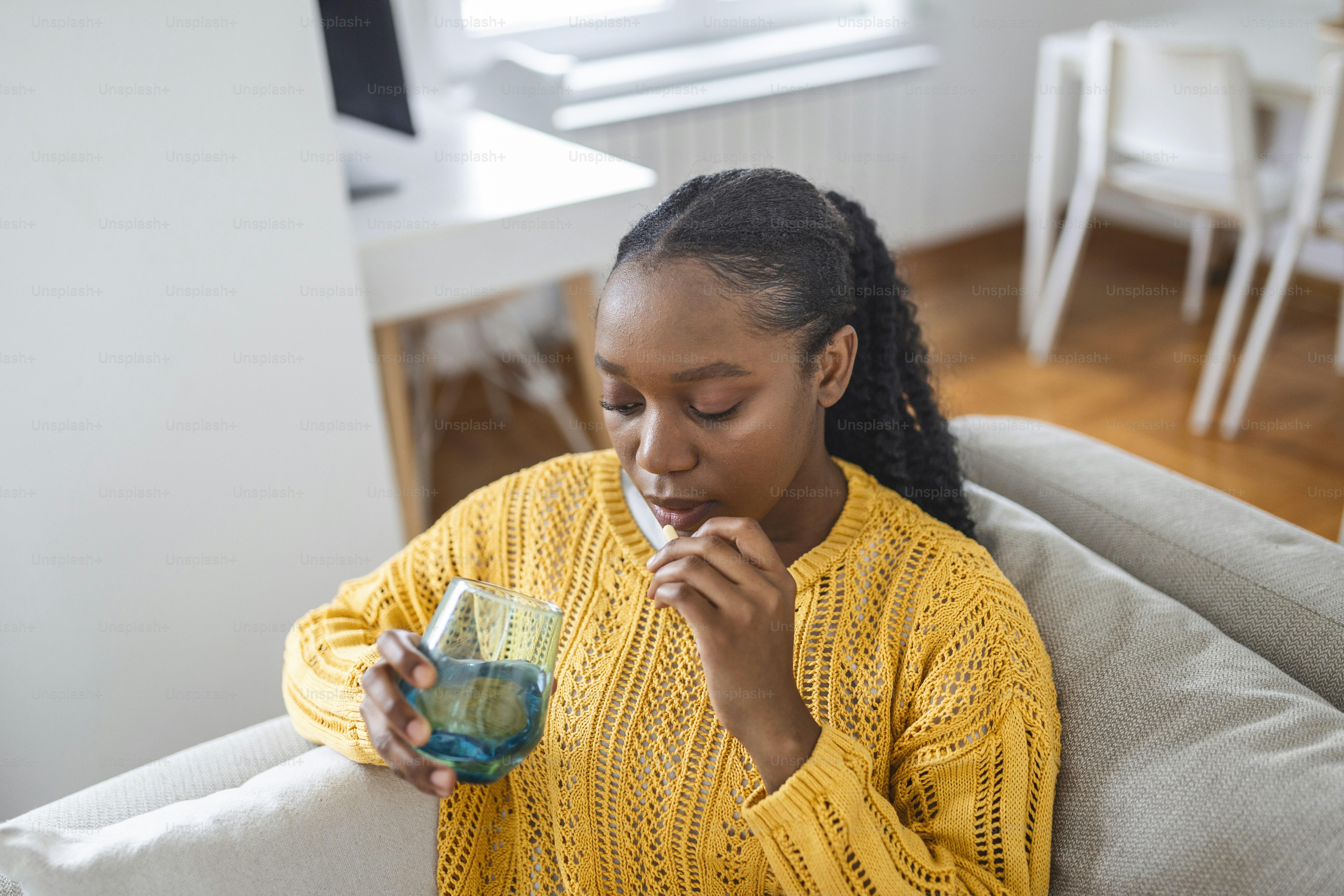 Concept of suffering from terrible pain. Beautiful, sad, upset, unhappy, troubled, weak woman with black hair, wearing casual clothes is sitting on a sofa and taking a pill