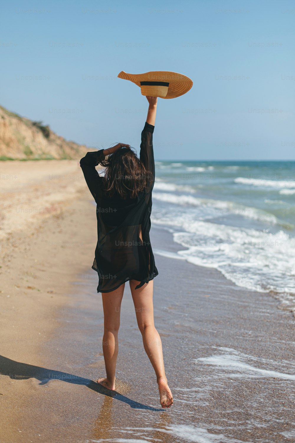 Belle femme insouciante avec chapeau marchant sur la plage de sable à la mer vagues et se relaxant. Vacances. Jeune femme élégante en chemise noire légère et chapeau de paille profitant de vacances sur une île tropicale