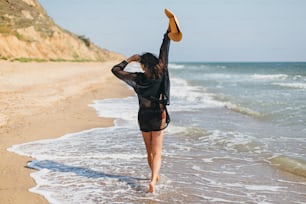 Summer vacation. Beautiful stylish woman with hat walking on sandy beach at sea waves and relaxing. Fit young female in light black shirt and straw hat enjoying vacation on tropical island. Carefree