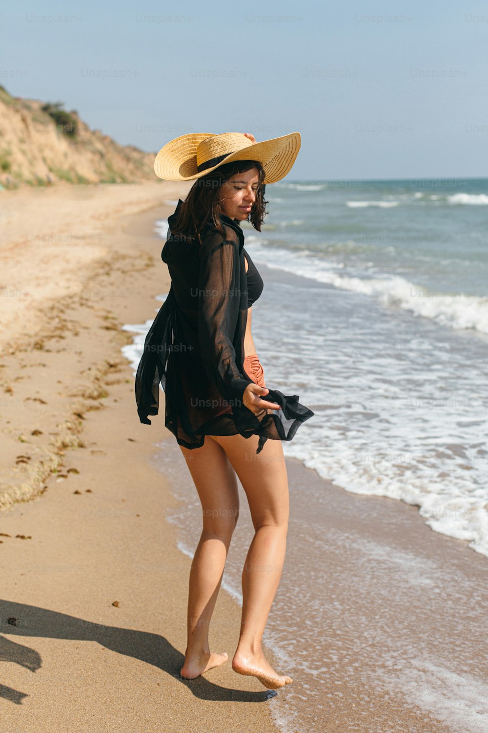 Mulher despreocupada bonita com chapéu andando na praia de areia nas ondas do mar e relaxante. Férias de verão. Fit elegante jovem fêmea na camisa preta clara e chapéu de palha desfrutando de férias na ilha tropical