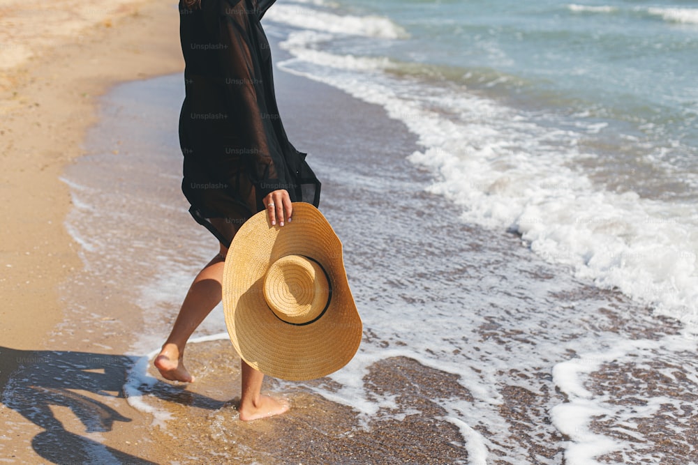 Carefree woman holding hat and walking on sunny beach at sea waves, close up. Summer vacation. Stylish young fit female with straw hat in hands  relaxing on tropical shore. Space for text