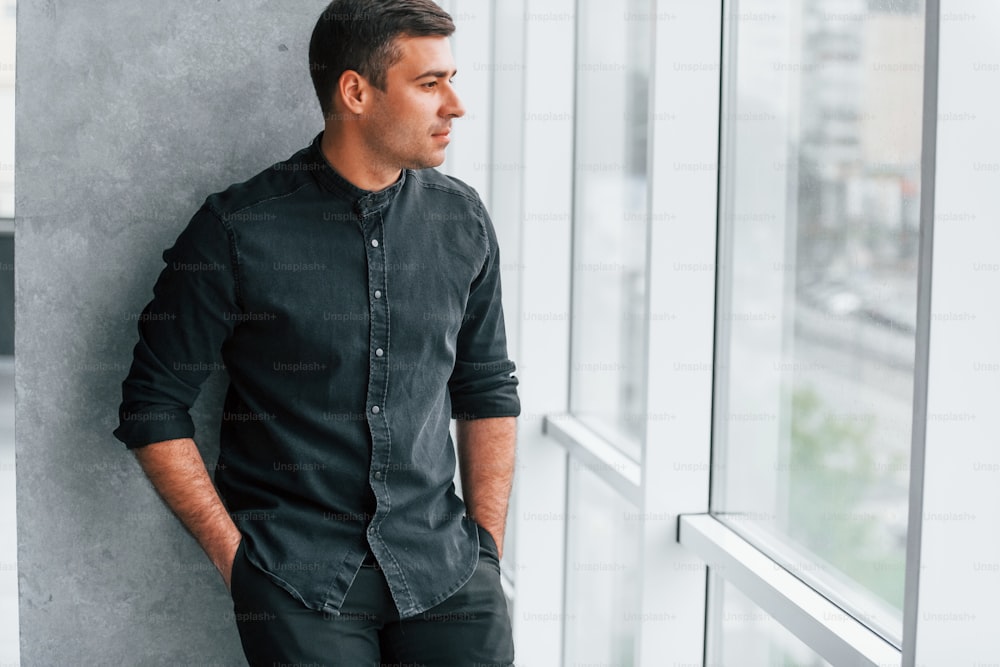 Elegant man in black shirt leaning on the wall indoors and looking through the big window.