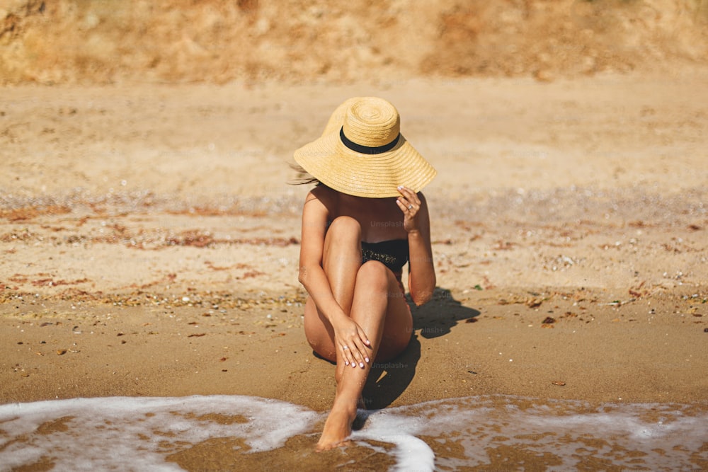 Beautiful stylish woman in hat sitting on sandy beach at sea waves and sunbathing. Summer vacation. Fit sexy young female in swimsuit and straw hat relaxing on tropical island. Space for text