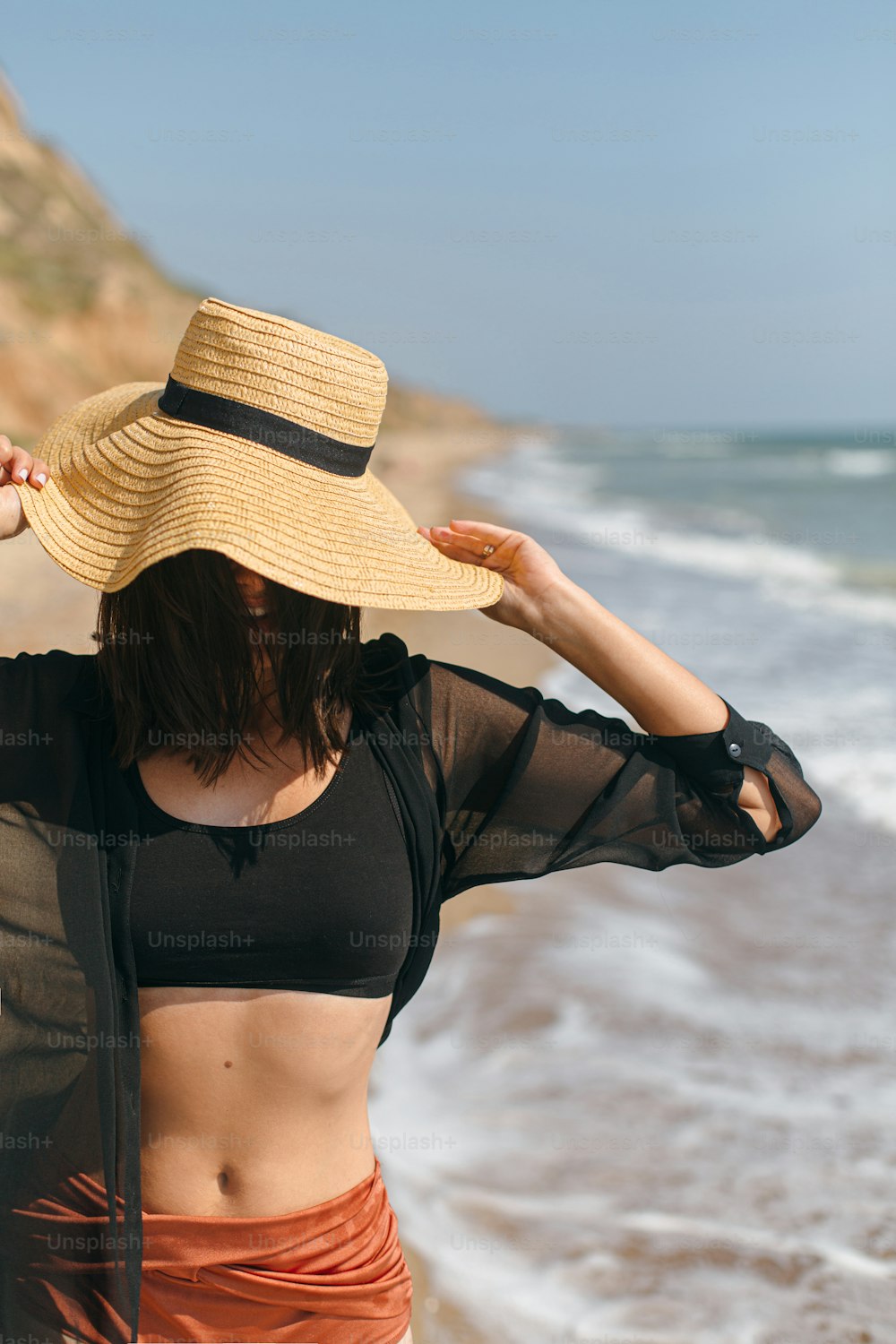 Stylish carefree woman in hat walking on sunny beach at sea and enjoying summer vacation. Young female in straw hat and light shirt relaxing on tropical island. Summer vacation
