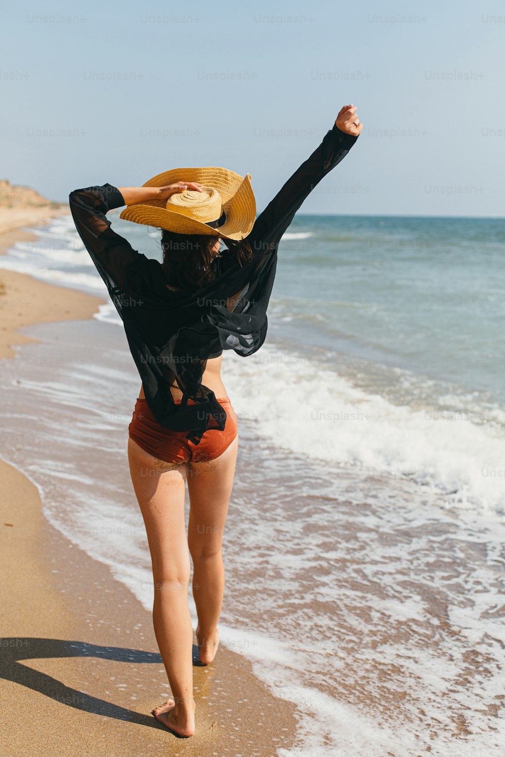 Belle femme insouciante avec chapeau marchant sur la plage de sable à la mer vagues et se relaxant. Vacances. Jeune femme élégante en chemise noire légère et chapeau de paille profitant de vacances sur une île tropicale
