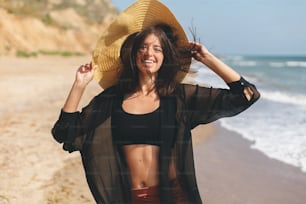 Beautiful happy woman with hat and windy hair relaxing and smiling on sandy beach at sea. Summer vacation.Stylish fit young female in light shirt and straw hat relaxing on tropical island