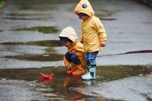 黄色い防水マントとブーツを身に着けた2人の子供が、雨上がりに屋外で手作りの紙のボートで遊んでいます。
