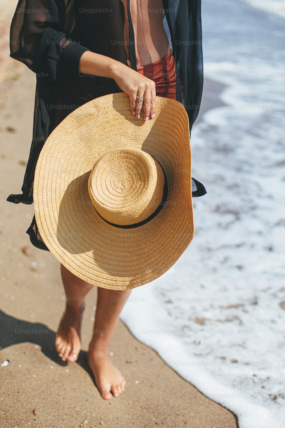 여름 방학. 햇볕이 잘 드는 해변에서 바다 파도를 걷고 있는 평온한 여자의 손에 세련된 모자가 닫힙니다. 밀짚 모자를 쓴 젊은 유행 여성은 열대 해안에서 휴식을 취하고 있습니다. 평온한