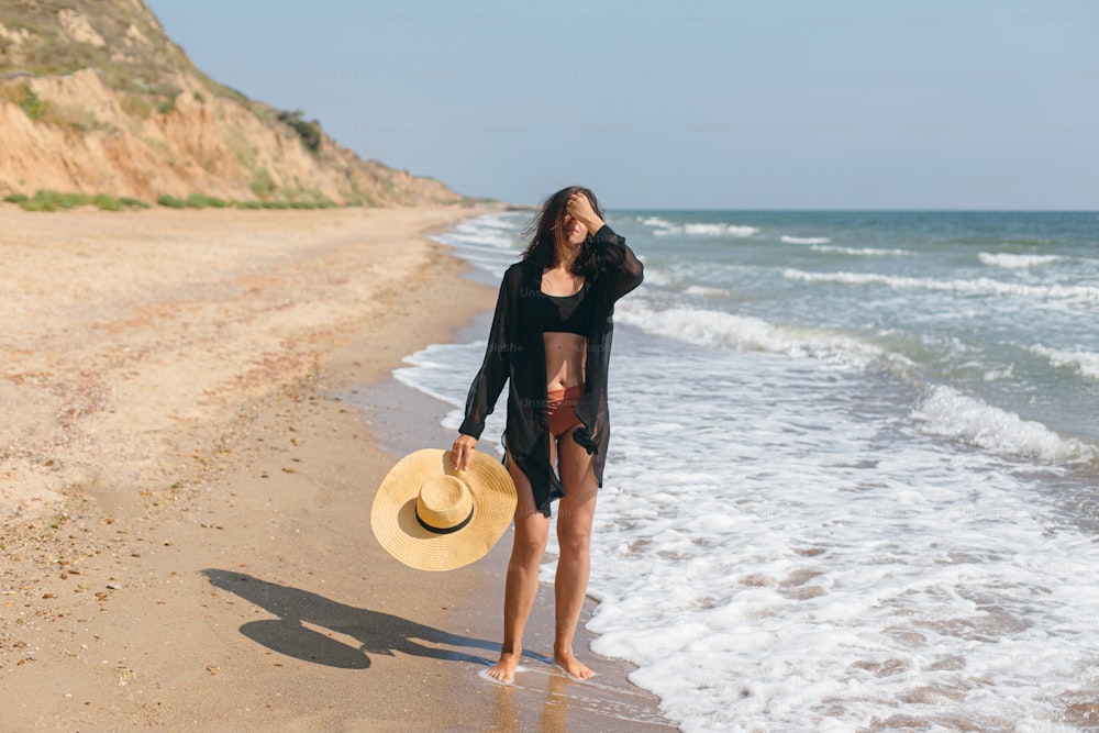 Beautiful carefree woman with hat walking on sandy beach at sea waves and relaxing. Summer vacation. Fit stylish young female in light black shirt and straw hat enjoying vacation on tropical island