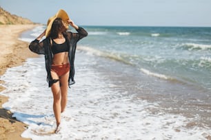 Beautiful carefree woman walking on sandy beach at sea waves and relaxing. Summer vacation. Stylish young female in light black shirt and straw hat enjoying tropical vacation. Space for text