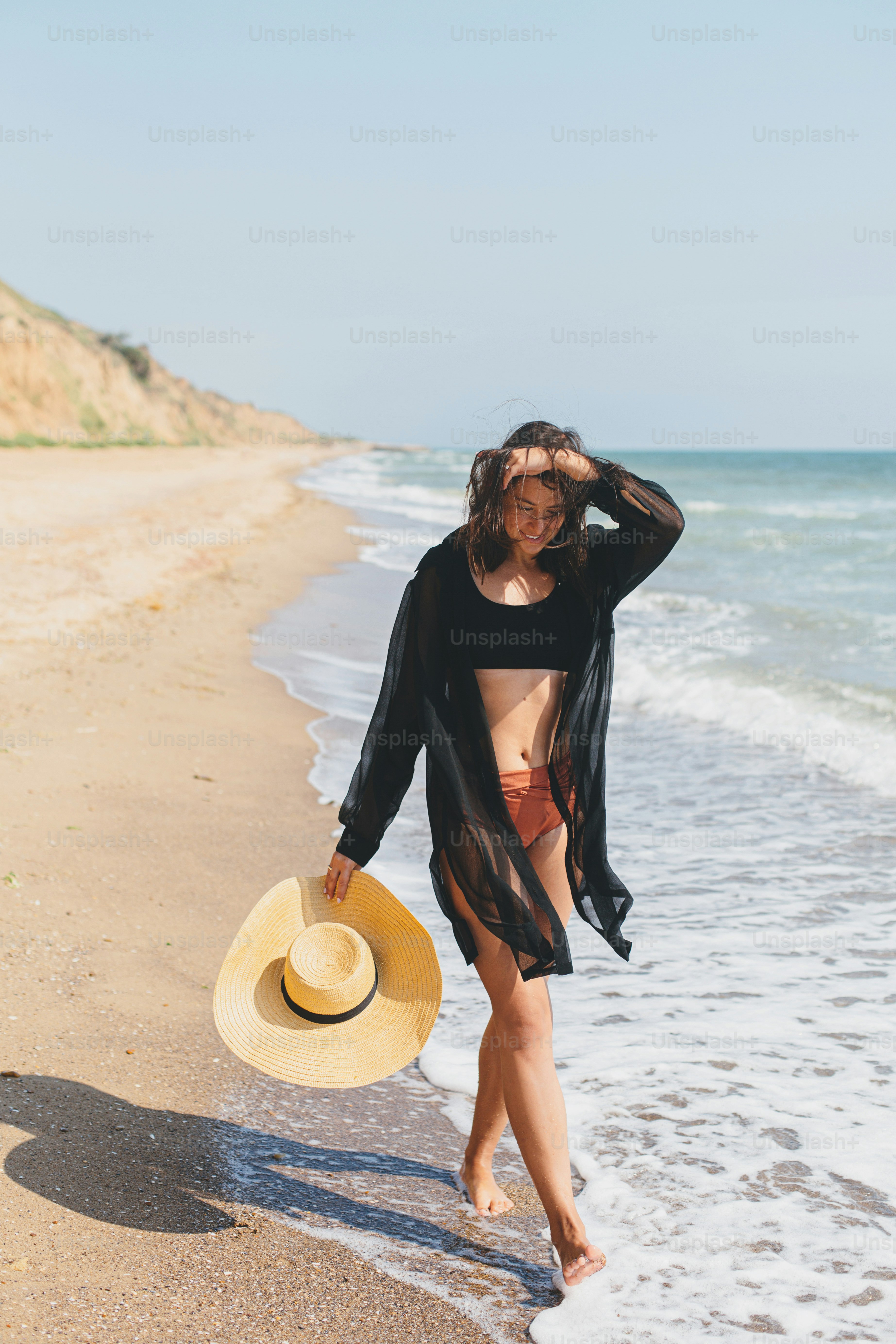 Beautiful carefree woman with hat walking on sandy beach at sea waves and relaxing. Summer vacation. Fit stylish young female in light black shirt and straw hat enjoying vacation on tropical island