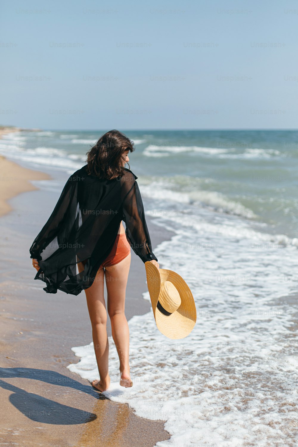Beautiful carefree woman with hat walking on sandy beach at sea waves and relaxing. Summer vacation. Fit stylish young female in light black shirt and straw hat enjoying vacation on tropical island
