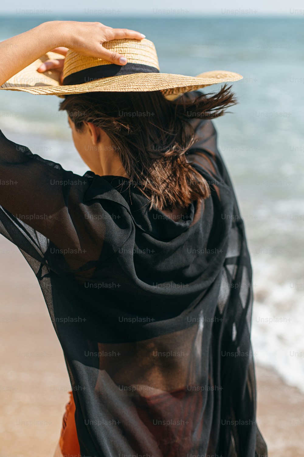 Beautiful stylish woman in hat walking on windy beach at sea waves. Summer vacation. Fit sexy young female in light shirt and straw hat relaxing on tropical island